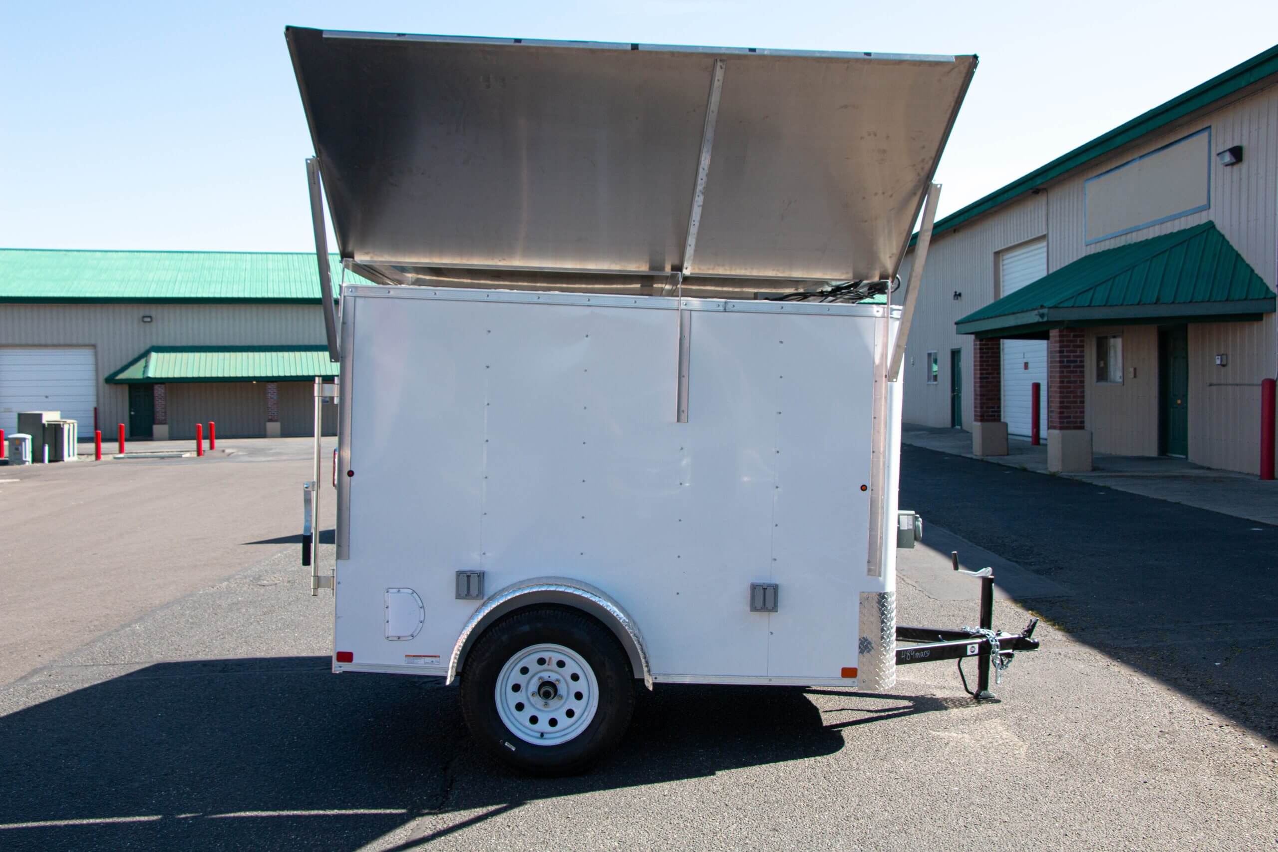 White utility trailer with an open hinged roof, parked on a paved lot near buildings with green roofs.
