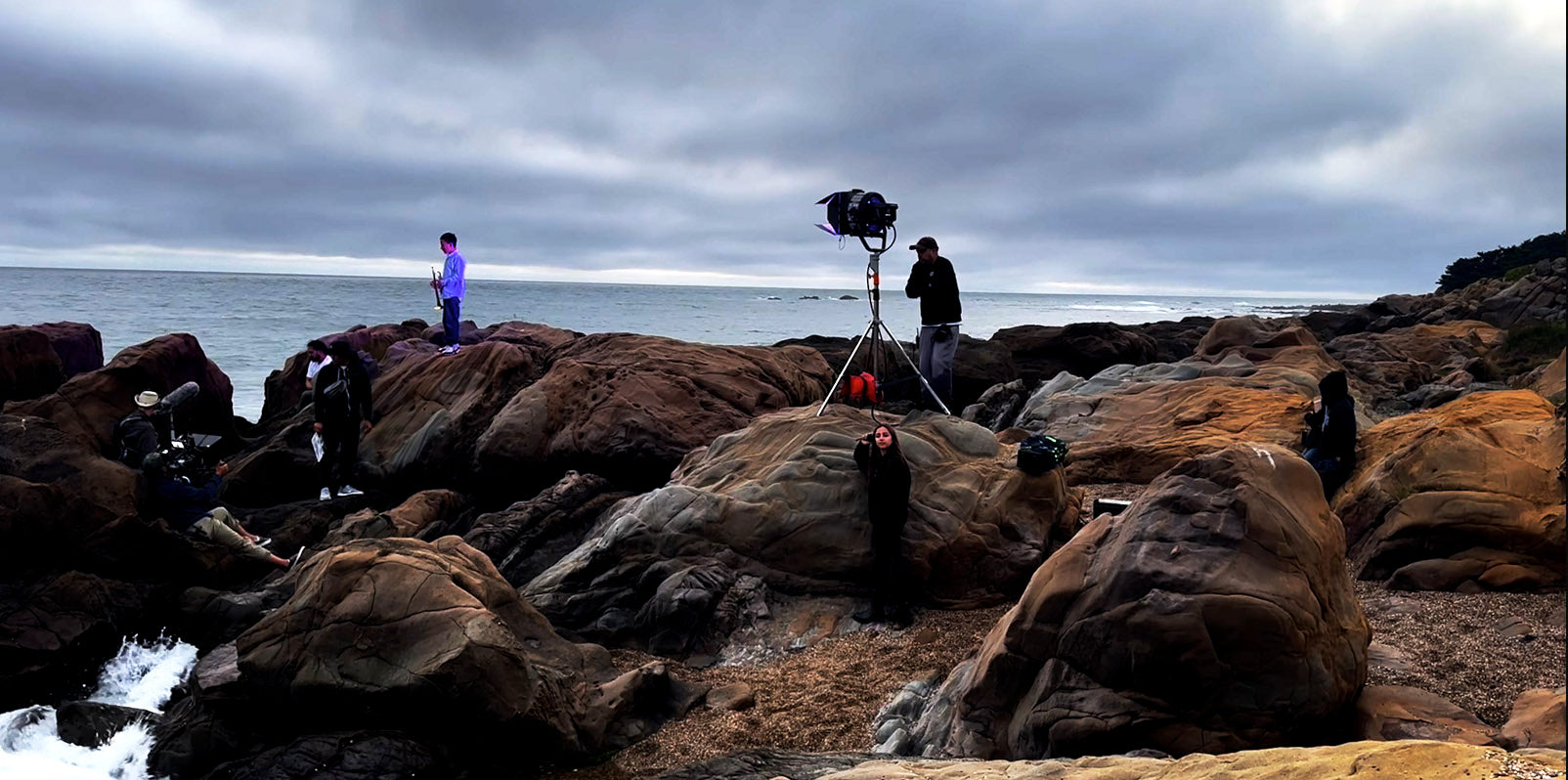 Film crew sets up equipment on rocky shoreline under cloudy skies, with one person standing near the edge overlooking the ocean.
