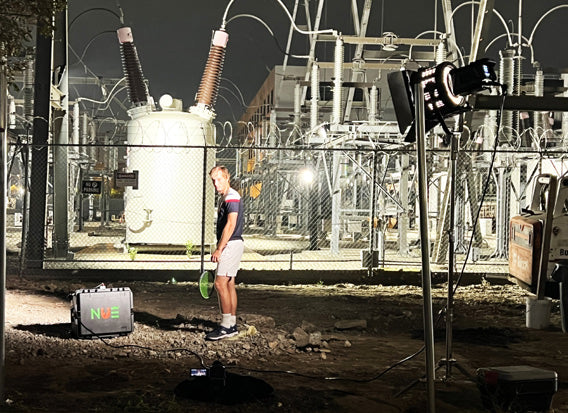Man stands holding a racket near a power substation at night, illuminated by lights and a portable NUE power case.