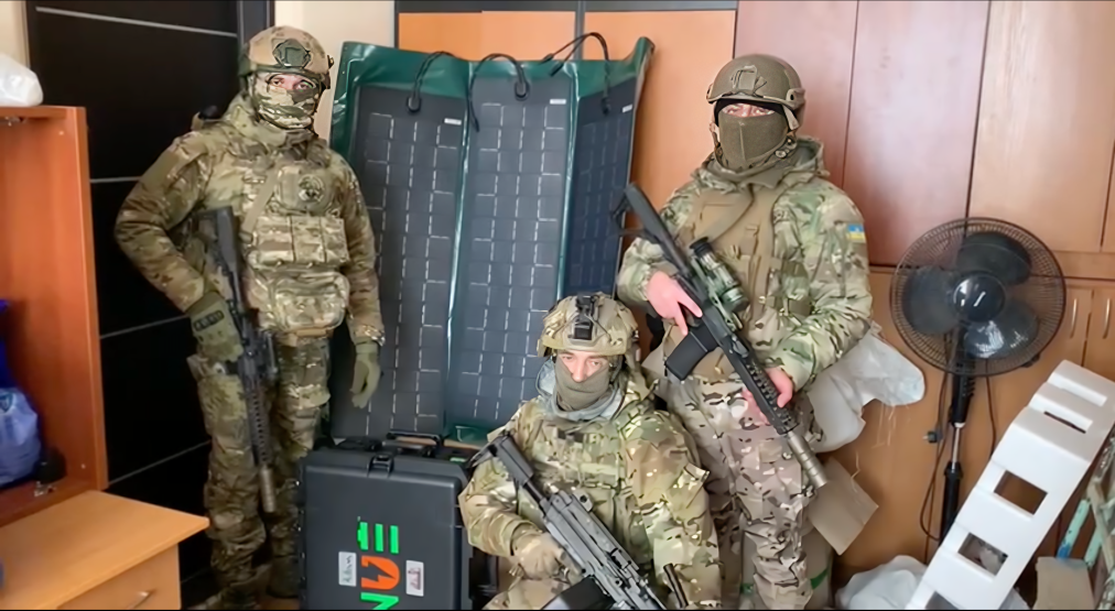 Three soldiers in camouflage gear posing in a room with a NUEPower battery