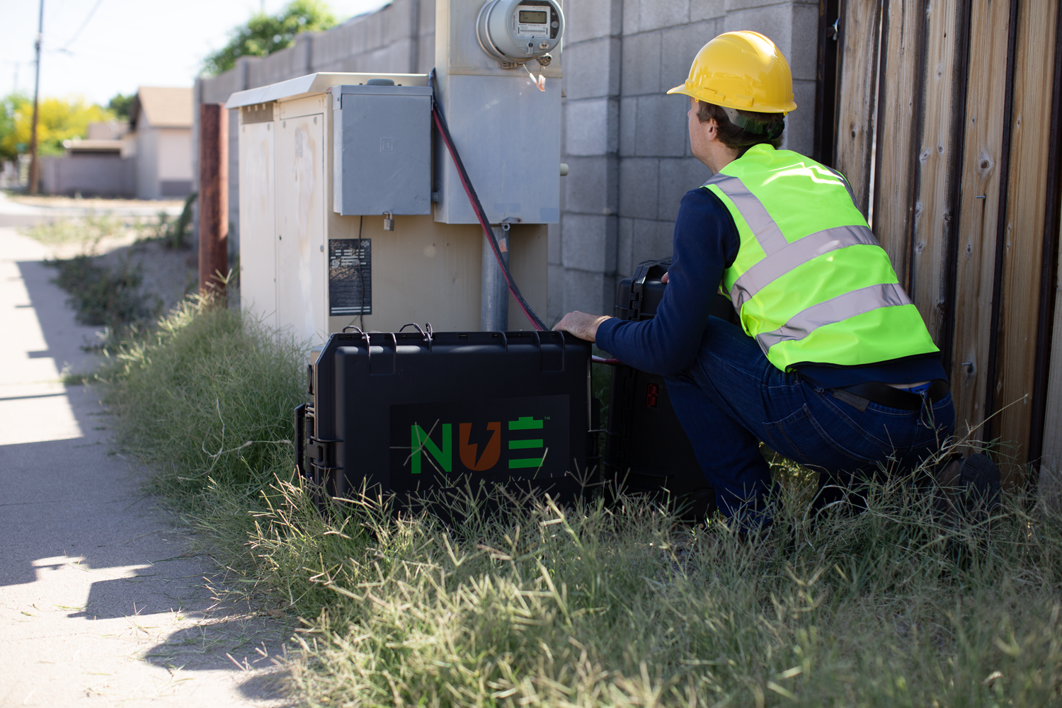 Worker in a yellow hard hat and safety vest installs a black NUE power system near an outdoor utility meter.